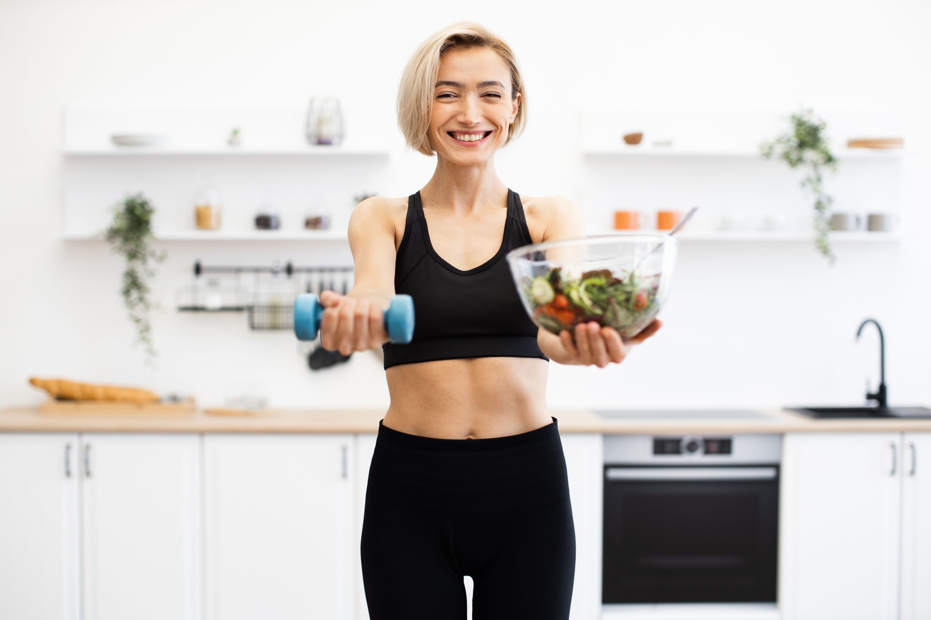 Smiling woman holding salad bowl and dumbbell in kitchen